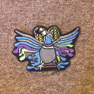 May include: A colorful embroidered patch featuring a stylized bird with outstretched wings. The bird is depicted in shades of blue, green, purple, and gray, with intricate details in yellow and black. The patch is perfect for adding a unique touch to clothing, bags, or other accessories.