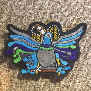 May include: A colorful embroidered patch featuring a stylized bird with blue, green, and purple wings. The bird is perched on a branch with a yellow and brown object behind it.