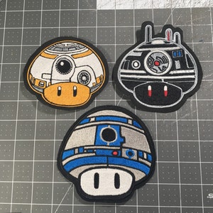 May include: Three embroidered iron-on patches featuring characters from Star Wars. The patches are shaped like mushrooms and feature a black and white R2-D2, a black and white Imperial Probe Droid, and a yellow and orange BB-8.