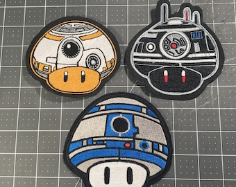 May include: Three embroidered iron-on patches featuring characters from Star Wars. The patches are shaped like mushrooms and feature a black and white R2-D2, a black and white Imperial Probe Droid, and a yellow and orange BB-8.