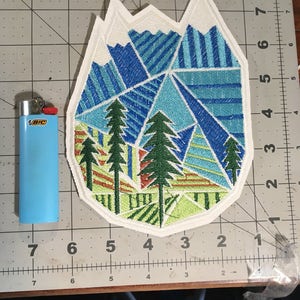 May include: White embroidered patch with a geometric design of blue, green, and yellow mountains and trees. The patch is shaped like a mountain peak.