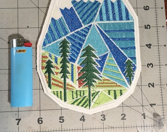 May include: White embroidered patch with a geometric design of blue, green, and yellow mountains and trees. The patch is shaped like a mountain peak.