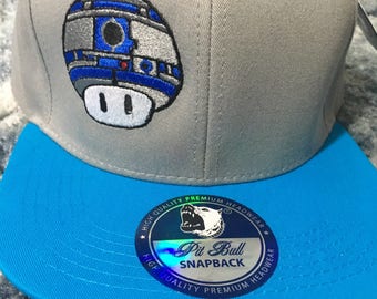 May include: Gray and blue baseball cap with a white and blue embroidered R2-D2 character on the front. The cap has a blue brim and a black and white sticker on the back that says "Pit Bull Snapback High Quality Premium Headwear".