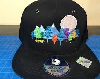 May include: Black baseball cap with a colorful embroidered design of a forest scene with mountains and a full moon. The cap has a white label with the text "Pit Bull Snapback" and a black and white logo of a pit bull.