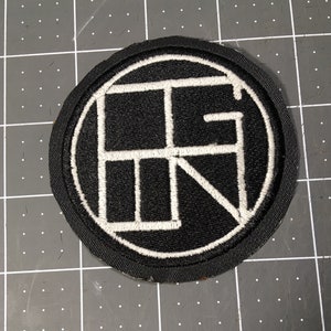 May include: Black and white embroidered patch with a circle design and the letter "R" inside. The patch is sewn onto a black fabric background.