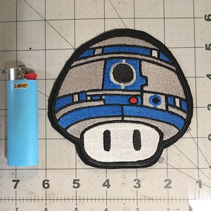 May include: A blue and gray embroidered patch of a mushroom with a R2-D2 design. The mushroom has a white stem with two black eyes.