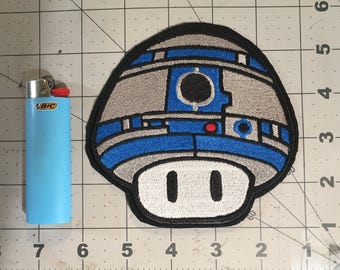 May include: A blue and gray embroidered patch of a mushroom with a R2-D2 design. The mushroom has a white stem with two black eyes.