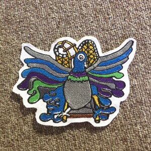 May include: Embroidered patch featuring a stylized pigeon with outstretched wings. The bird has a gray body, blue head, and wings in shades of blue, purple, and green. The patch includes yellow and brown accents and a white border.
