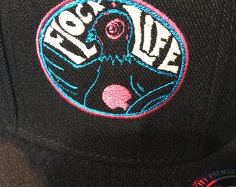 May include: Black baseball cap with an embroidered patch. The patch features the words "FLOCK LIFE" in white, surrounding a stylized figure with a pink heart and a swirling eye. The patch is outlined in blue and pink.
