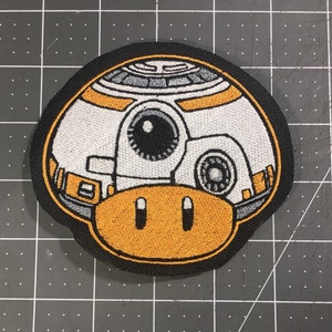 May include: Embroidered patch of a white and orange robot with a black eye, a black and gray circular detail, and a yellow bottom. The robot is on a black background.