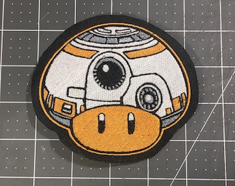 May include: Embroidered patch of a white and orange robot with a black eye, a black and gray circular detail, and a yellow bottom. The robot is on a black background.