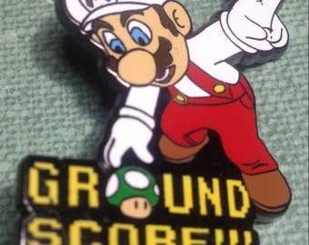 May include: A red and white enamel pin featuring a cartoon character of Mario from the Super Mario video game series. Mario is wearing his signature red overalls and white gloves. He is holding a green mushroom and giving a peace sign. The text "GROUND SCORE!!" is written in a pixelated font below Mario.