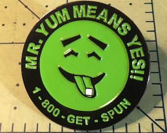 May include: A black and green enamel pin with a smiling face and the text "Mr. Yum Means Yes! 1-800-Get-Spun"