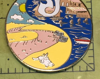 May include: A circular pin with a yin yang design. The left side is yellow with a pink and white cow skull, a sun, and clouds. The right side is blue with a white building, a white boat, and a white 'SJ' with a smiley face.