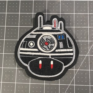 May include: Black and white embroidered patch of a robot with red and blue details. The robot has a round body with a large central eye and two smaller eyes on the top. The robot has two antennas on top of its head and two small legs on the bottom.