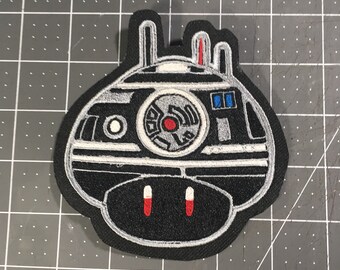 May include: Black and white embroidered patch of a robot with red and blue details. The robot has a round body with a large central eye and two smaller eyes on the top. The robot has two antennas on top of its head and two small legs on the bottom.