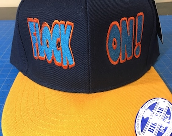 May include: A navy blue and yellow snapback hat with the text "FLOCK ON!" in blue and orange embroidery. The hat has a flat brim and a Big Gear Snapback label.