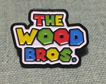 May include: A black and white enamel pin with the text "THE WOOD BROS." in colorful letters. The letters are outlined in black and filled with different colors: yellow, green, red, and blue.