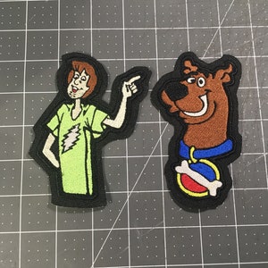May include: Two embroidered patches featuring characters from the cartoon Scooby-Doo. The first patch is green and features Shaggy with brown hair and a green shirt. The second patch is brown and features Scooby-Doo with a blue collar and a red and white bone.