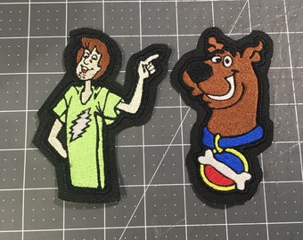 May include: Two embroidered patches featuring characters from the cartoon Scooby-Doo. The first patch is green and features Shaggy with brown hair and a green shirt. The second patch is brown and features Scooby-Doo with a blue collar and a red and white bone.