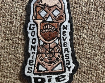 May include: A black and white embroidered patch with a skull and crossbones design. The skull is brown and has a white face with a black crossbones. The text "Goonies Never Say Die" is written around the skull in white.
