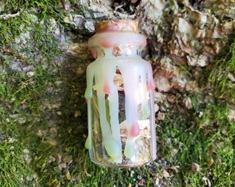 May include: A small glass bottle with a cork stopper is filled with dried herbs and sealed with a layer of melted wax. The wax is a light green and pink color and has dripped down the sides of the bottle.