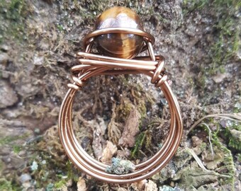 May include: A copper wire wrapped ring with a tiger's eye gemstone. The ring is set against a mossy background.