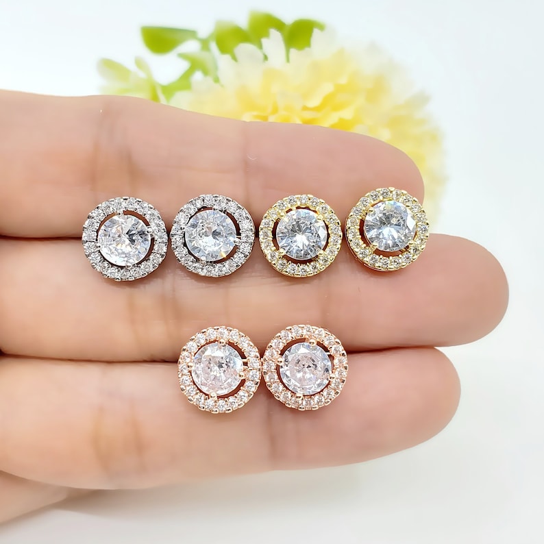 May include: Three pairs of round stud earrings with a halo of smaller stones surrounding a larger center stone. The earrings are shown in silver, gold, and rose gold.