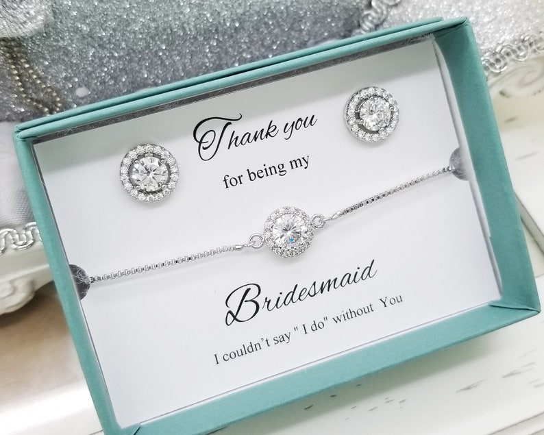 May include: A white gift box with a teal lid, containing a silver bracelet and earrings with clear stones. The box has a message that reads "Thank you for being my Bridesmaid. I couldn't say "I do" without you."