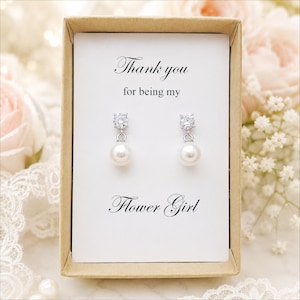 May include: A pair of silver-toned drop earrings featuring a clear round stone stud and a white pearl dangle, presented in a tan cardboard box with a white card printed with the text Thank you for being my Flower Girl in black script.