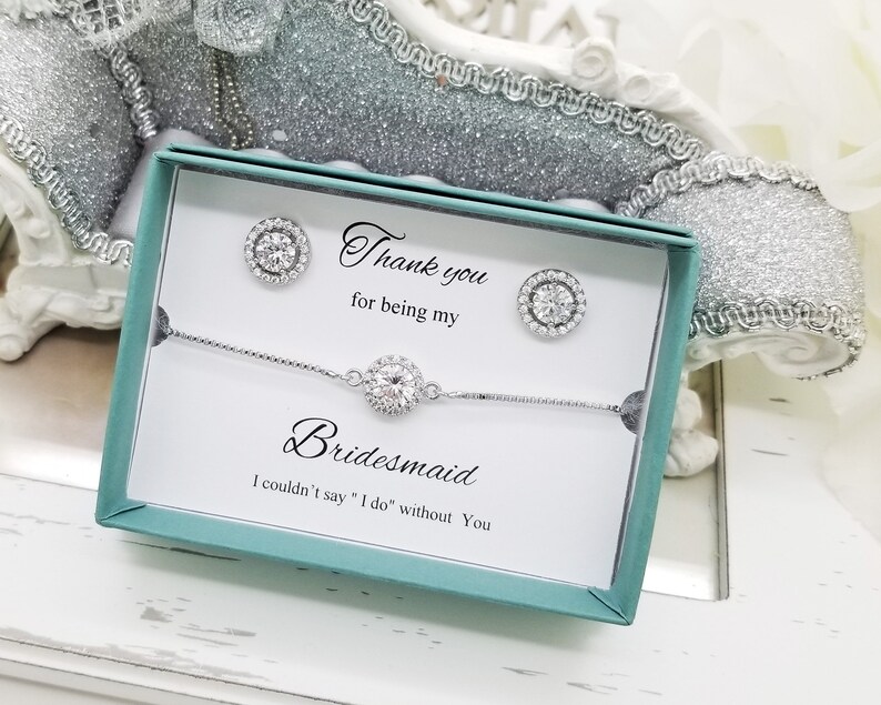 May include: A teal gift box with a white card inside. The card says "Thank you for being my Bridesmaid. I couldn't say "I do" without you." The box contains a silver bracelet and a pair of silver earrings with clear stones.