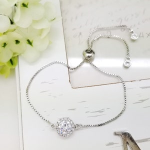 May include: A silver adjustable bracelet with a round, clear crystal centerpiece. The crystal is surrounded by smaller crystals in a halo design.