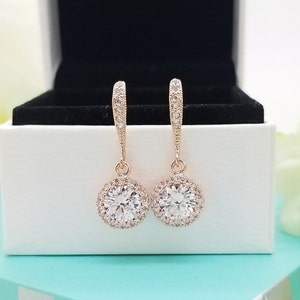 May include: A pair of rose gold dangle earrings with a round, clear crystal stone surrounded by smaller crystals. The earrings have a simple, elegant design.