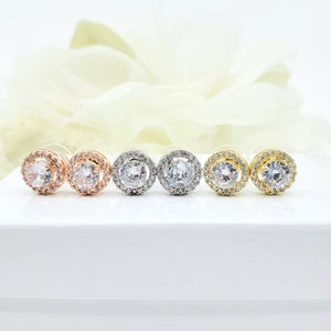 May include: Four pairs of round halo stud earrings in silver, rose gold, and gold finishes. Each earring features a clear round cubic zirconia stone surrounded by a halo of smaller cubic zirconia stones.