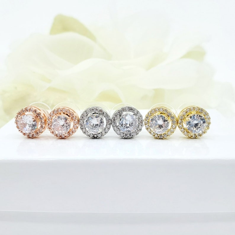May include: Four pairs of round halo stud earrings in silver, rose gold, and gold finishes. Each earring features a clear round cubic zirconia stone surrounded by a halo of smaller cubic zirconia stones.