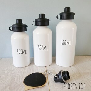 Peut inclure: Trois gourdes blanches avec des bouchons noirs, &eacute;tiquet&eacute;es 400ml, 500ml et 600ml. Un bouchon noir avec un porte-cl&eacute;s et un bouchon noir s&eacute;par&eacute; sont &eacute;galement pr&eacute;sent&eacute;s. Le texte "SPORTS TOP" est visible.