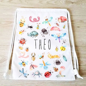 May include: White drawstring bag decorated with watercolour illustrations of insects such as bees, ladybirds, and dragonflies. The name "THEO" is printed in black. The bag has white drawstrings.