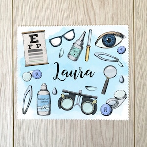 Peut inclure: Un fond bleu clair avec des illustrations d'outils d'optométrie, dont un tableau de lecture, des lunettes, des gouttes pour les yeux et des lentilles de contact. Le nom "Laura" est écrit en cursive. Le design est sur fond blanc à bord festonné.