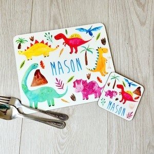 Peut inclure: Un set de table et un dessous de verre blancs avec des dinosaures colorés à l'aquarelle, des palmiers et le nom "MASON" en bleu. Le set est conçu pour les enfants. Un ajout amusant et coloré à toute table.