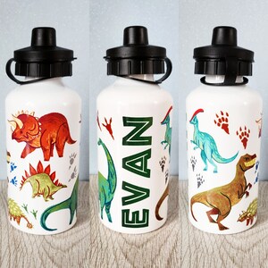 Peut inclure: Gourde blanche avec un bouchon noir, orn&eacute;e d'illustrations color&eacute;es de dinosaures et du nom "EVAN" en vert. Les dinosaures incluent un Tric&eacute;ratops, un St&eacute;gosaure et un T-Rex, avec des empreintes de pas de dinosaures.