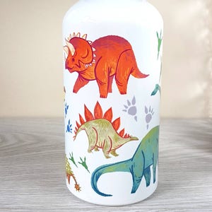 Peut inclure: Gourde blanche orn&eacute;e d'illustrations color&eacute;es de dinosaures, dont un Tric&eacute;ratops, un St&eacute;gosaure et un Brontosaure. Le motif comprend des empreintes de pas de dinosaures bleues et grises, avec des accents de feuillage vert. La gourde est cylindrique.