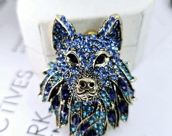 Könnte beinhalten: Eine blaue und goldene Wolfskopf-Brosche mit Strasssteinen. Die Augen des Wolfes sind schwarz und die Nase ist golden. Die Brosche ist ein detailliertes und kunstvolles Schmuckstück.