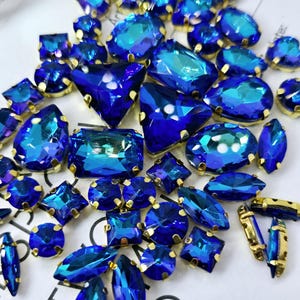 Può includere: Assortimento di strass di vetro blu iridescente con montature dorate. Gli strass sono di varie forme e dimensioni, tra cui rotondi, ovali, quadrati e a goccia.