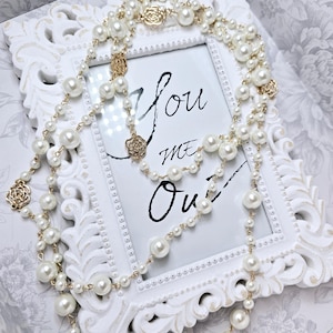 May include: Elegant pearl necklaces with gold accents are draped over a white framed picture. The picture has the words "You me Oui" written on it. The necklaces feature various sizes of faux pearls and gold rose-shaped charms.