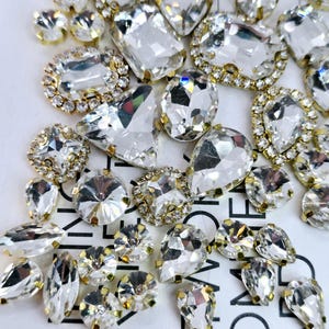 May include: Assortment of clear glass rhinestones with gold settings. The rhinestones are in various shapes and sizes, including round, oval, teardrop, and square.