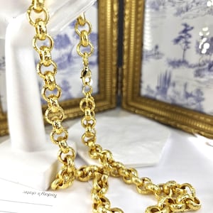 May include: A gold-tone chain necklace featuring a series of interlocking circular links with a textured, hammered finish. The necklace includes a secure lobster claw clasp and is displayed on a white stand, showcasing the metallic lustre and design.