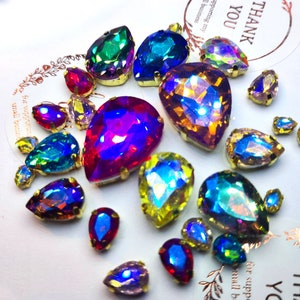 May include: Assortment of iridescent teardrop-shaped rhinestones with gold settings. The rhinestones are in various colours, including green, blue, purple, red, and yellow.