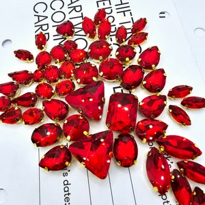 May include: A selection of red gemstones in different shapes, such as teardrops, ovals, and squares, each with a gold-coloured metal setting. The gemstones are arranged on a white surface with printed text.