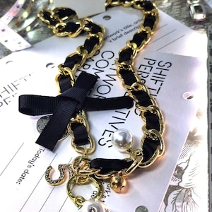 May include: Gold-tone chain necklace with black ribbon detailing. Includes a black bow, pearl and rhinestone charms, and a small gold bell. The necklace has the text "Fashion Jewelry" on a tag.