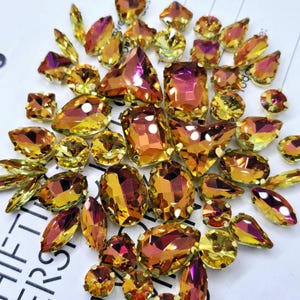May include: Assortment of iridescent glass rhinestones in various shapes and sizes. The rhinestones have a mix of gold and pink hues.